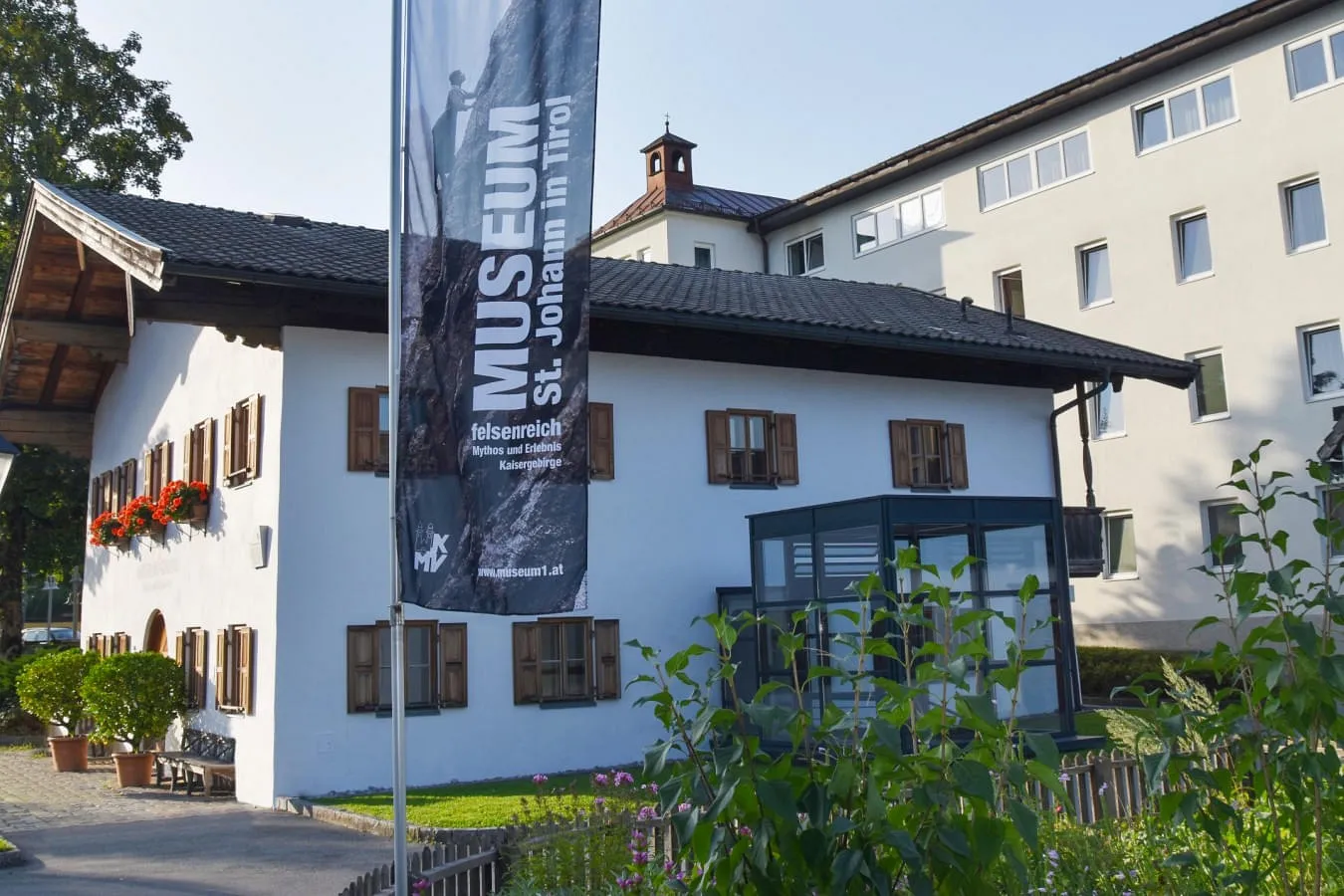 Außenansicht des Museums St. Johann in Tirol, mit einer Fahne, die das Museum bewirbt. Das Gebäude hat eine weiße Fassade mit braunen Fensterläden und Blumenkästen. 
